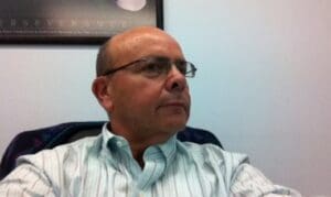 A man with glasses sitting in front of a wall.