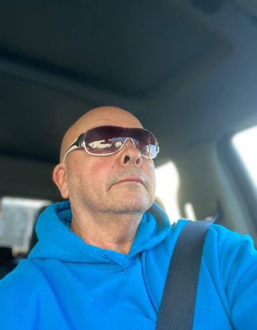 A bald man wearing sunglasses and blue hoodie.